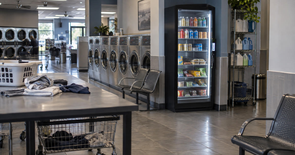 Revvolto smart fridge in a laundromat waiting area with washers, folding tables, and clear service access
